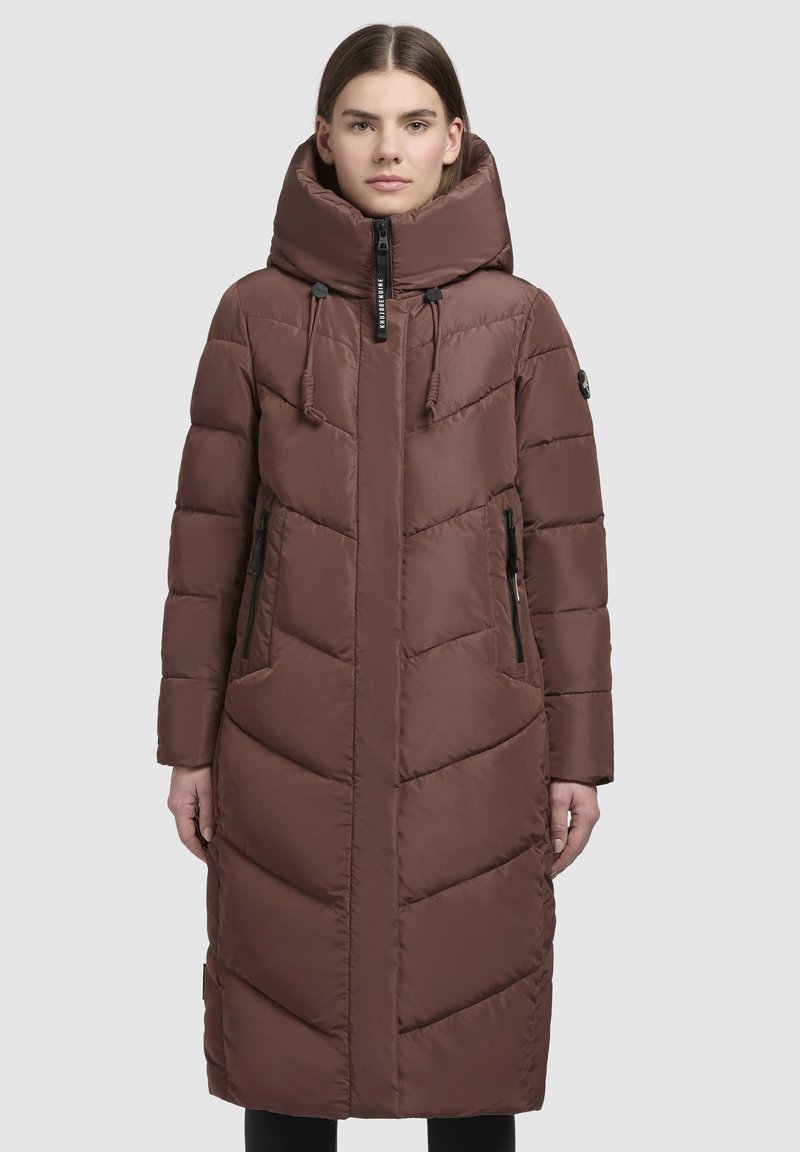 Kvindelig iført en lang, brun quilted pufferjakke med høj krave, justerbar hætte og lynlåslommer på siden, stående foran en ensfarvet baggrund.