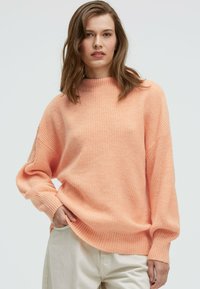 Oversized sweater in soft peach knit, featuring a high neckline and ribbed texture, paired with light-colored pants.