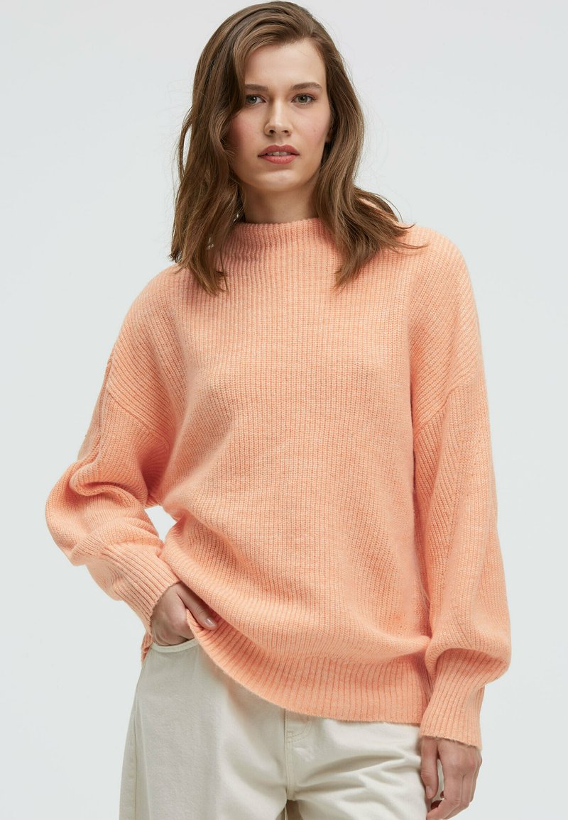 Oversized sweater in soft peach knit, featuring a high neckline and ribbed texture, paired with light-colored pants.