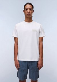 Balta īsroku T-krekls ar apaļu izgriezumu. Uz krūtīm ir taisnstūra formas uzlīme, un materiālam ir gluda kokvilnas tekstūra. Valdāts kopā ar ziliem šortiem.