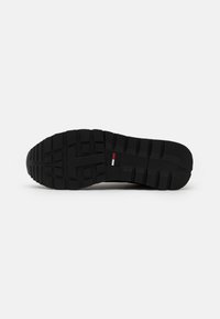 Semelle de chaussure noire avec des rainures profondes pour la traction et un petit logo rectangulaire rouge, blanc et bleu au centre.