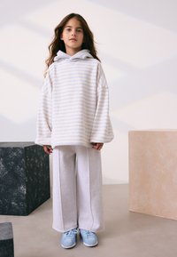 Hanorac supradimensionat cu dungi crem și glugă gri, asortat cu pantaloni sport largi gri deschis și adidași albaștri. Material neted și croială lejeră.