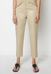 Pantalon beige, coupe ajustée, texture lisse, longueur raccourcie, passants pour ceinture, design classique, associé à des sandales noires à enfiler.