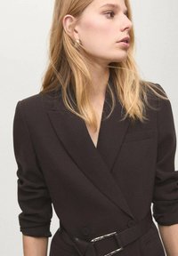 Ciemnobrązowa marynarka o dopasowanym kroju, z klapami z wycięciem, pojedynczą kieszenią, podwiniętymi rękawami oraz paskiem w talii z metalowymi zdobieniami.