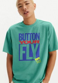 Teal cotton t-shirt with bold graphic text in red and blue; features "BUTTON YOUR FLY" and "Levi's 501" logo in yellow at the bottom.