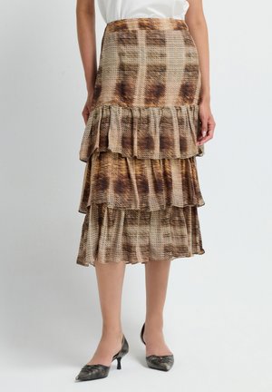 Donna che indossa una gonna a strati marrone e beige con motivo astratto e décolleté metallici a punta bassa su uno sfondo uniforme.