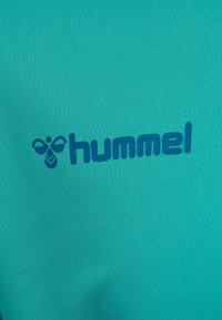 Teal Sporttrikot aus strukturiertem Stoff, mit einem blauen Logo, das eine stilisierte Biene und den markanten Markennamen „hummel“ zeigt.