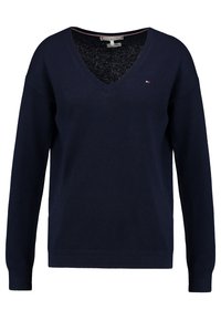 Tmavě modrý svetr s výstřihem do V vyrobený z měkké pleteniny. Má dlouhé rukávy a texturované manžety, s malým logem na hrudi.