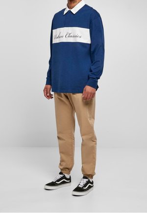 Marineblaues Langarmshirt mit weißem horizontalem Streifen und dem Schriftzug "Urban Classics"; kombiniert mit beigefarbenen Hosen und schwarz-weißen Turnschuhen.