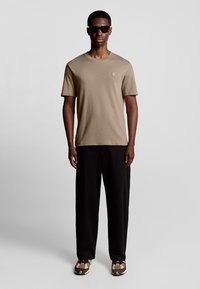 Beige T-Shirt mit Logo, schwarze weit geschnittene Hose und gemusterte braune Sneakers; das Model trägt dunkle Sonnenbrillen.