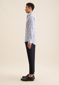 Langarmshirt in lichtblauw met wit bloemenpatroon, gecombineerd met zwarte tapered broeken en zwarte schoenen, bekeken vanuit de zijkant.