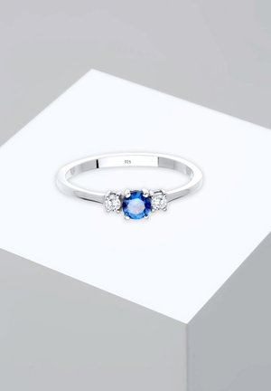 Bague en argent avec une pierre précieuse bleue centrale flanquée de deux pierres claires, monté sur un bandeau lisse avec le marquage "925" à l'intérieur.