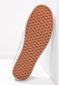 Semelle de chaussure avec un motif gaufré en caoutchouc marron, inclinée à la verticale contre un mur blanc et un sol.
