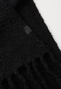 Écharpe noire texturée avec une surface douce et duveteuse et des bords à franges. Comprend une petite étiquette en tissu noir avec un texte en relief.
