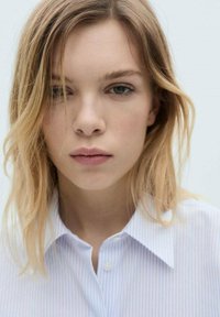 Collared shirt in light blue with thin white stripes. Model has wavy, shoulder-length blonde hair and a neutral expression.
