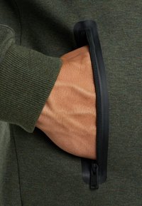 Zelená tkaninová mikina s kapucňou a čiernou zipsovou vreckom. Vrecko má hladký, gumovaný okraj. Textúra rebrovaného manžetu je viditeľná.
