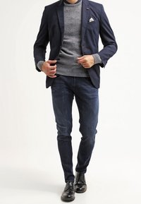 Blazer bleu marine sur un pull gris, accompagné d'un jean bleu et de chaussures noires. Détails notables comprenant un mouchoir de poche et un tricot texturé du pull.