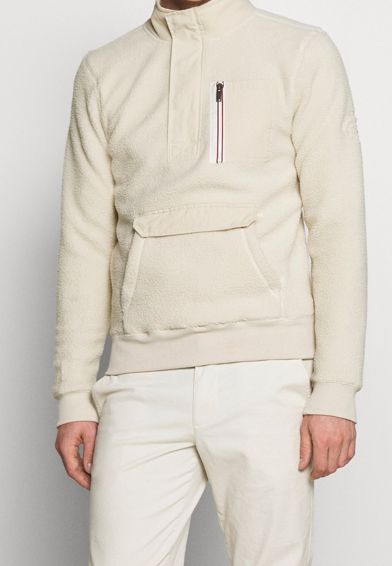 Pullover in pile color crema con collo alto, tasca sulla chest con zip, tasca a marsupio frontale e polsini e orlo a coste.