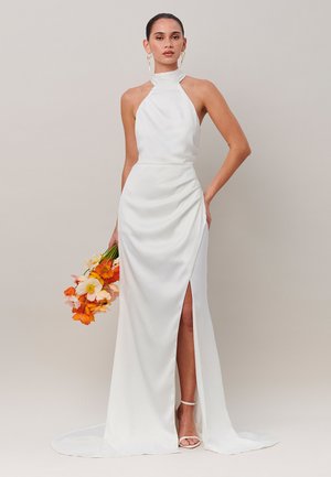 Vrouw in een witte halter-nekgordeljurk met een split aan de dij, houdt oranje en witte bloemen vast, draagt gouden oorbellen en witte hakken.