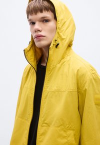Κίτρινο μπουφάν με κουκούλα, κλείσιμο με φερμουάρ και πλαϊνές τσέπες. Ελαφρύ υλικό με λεία υφή. Ετικέτα εμπορικού σήματος στο μανίκι.