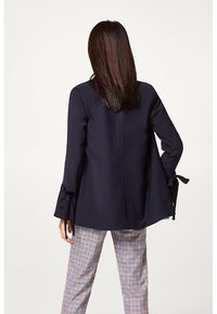 Nainen, joka on pukeutunut laivastonsiniseen bleiseriin, jonka hihnat on sidottu, ja harmaisiin ruudullisiin housuihin, seisoo selkä kameraan päin vaaleaa taustaa vasten.