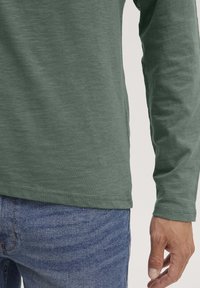 Langärmliges grünes T-Shirt aus strukturiertem Baumwollstoff, das einen lockeren Schnitt mit Rundhalsausschnitt und genähtem Saum aufweist, kombiniert mit blauen Jeans.