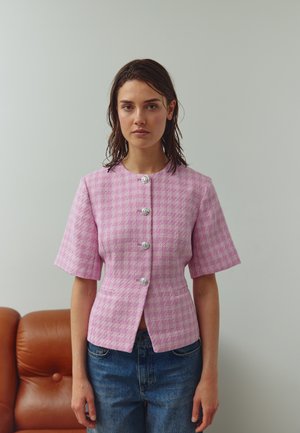 Ženska z mokrimi lasmi, oblečena v rožnato karo kratkoročni blazer s štirimi srebrnimi gumbi in modrimi jeans hlačami, stoji v notranjosti ob rjavi usnjeni sedežni garnituri.