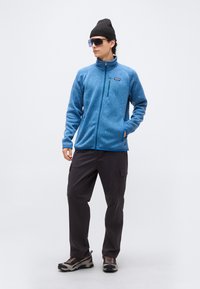 Άνδρας που φορά μπλε φλις μπουφάν με φερμουάρ, μαύρο παντελόνι καμπαρντίνα, παπούτσια πεζοπορίας, μαύρο σκουφί και μεγάλα γυαλιά ηλίου, στέκεται μπροστά σε λευκό φόντο.