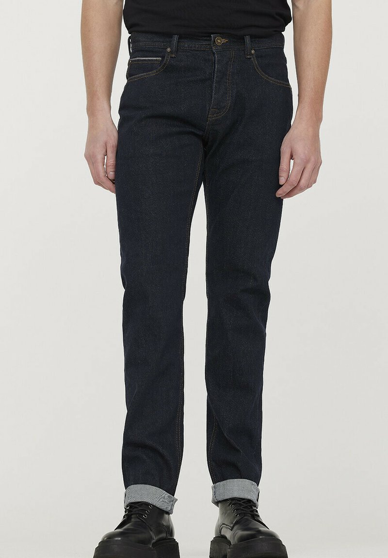 Jeans en denim bleu foncé à coupe slim, comportant cinq poches, des ourlets roulés bleu clair, et des coutures orange contrastantes.