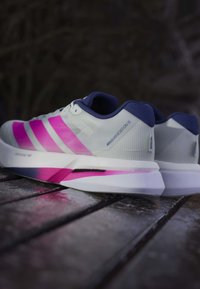 Paire de chaussures de course Adidas Adizero Boston 13 blanches et roses avec des accents bleu marine, posées sur une surface en bois à l'extérieur au crépuscule.