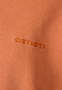 Narančasta pletenina s suptilnom teksturom, s izvezenim logotipom "Carhartt" u odgovarajućoj nijansi.