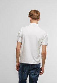 Balta īsām piedurknēm polo krekls ar klasisku apkakli, gluda tekstūra un smalkas izšuvumu detaļas, apvienots ar ziliem džinsu biksēm.