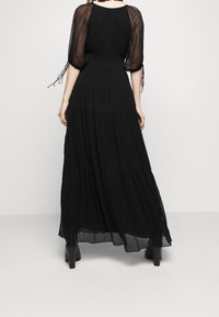 Czarna sukienka maxi z przezroczystymi, zwiewnymi rękawami i karbowanym gorsetem. Posiada warstwowe falbany i płynną sylwetkę. Noszona z botkami na obcasie.
