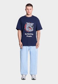 Granatowa koszulka bawełniana z grafiką szarego buldoga z czerwonymi akcentami i napisem "No Barking Prosto" na białym tle, noszona z jasnoniebieskimi dżinsami.