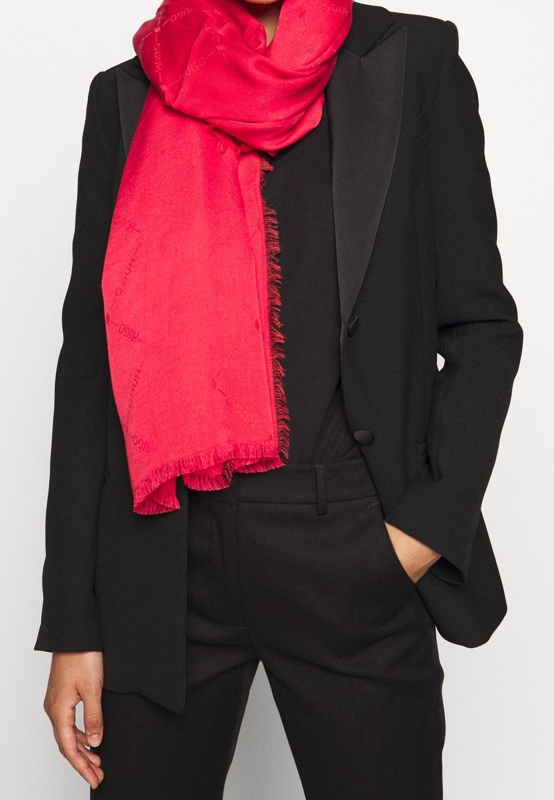 Personne portant un costume noir et une écharpe rouge à franges, une main dans la poche, sur un fond clair uni.