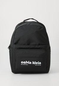 Sac à dos en nylon noir avec une forme arrondie, poche zippée à l'avant, marquage blanc proéminent et dos rembourré pour le confort.