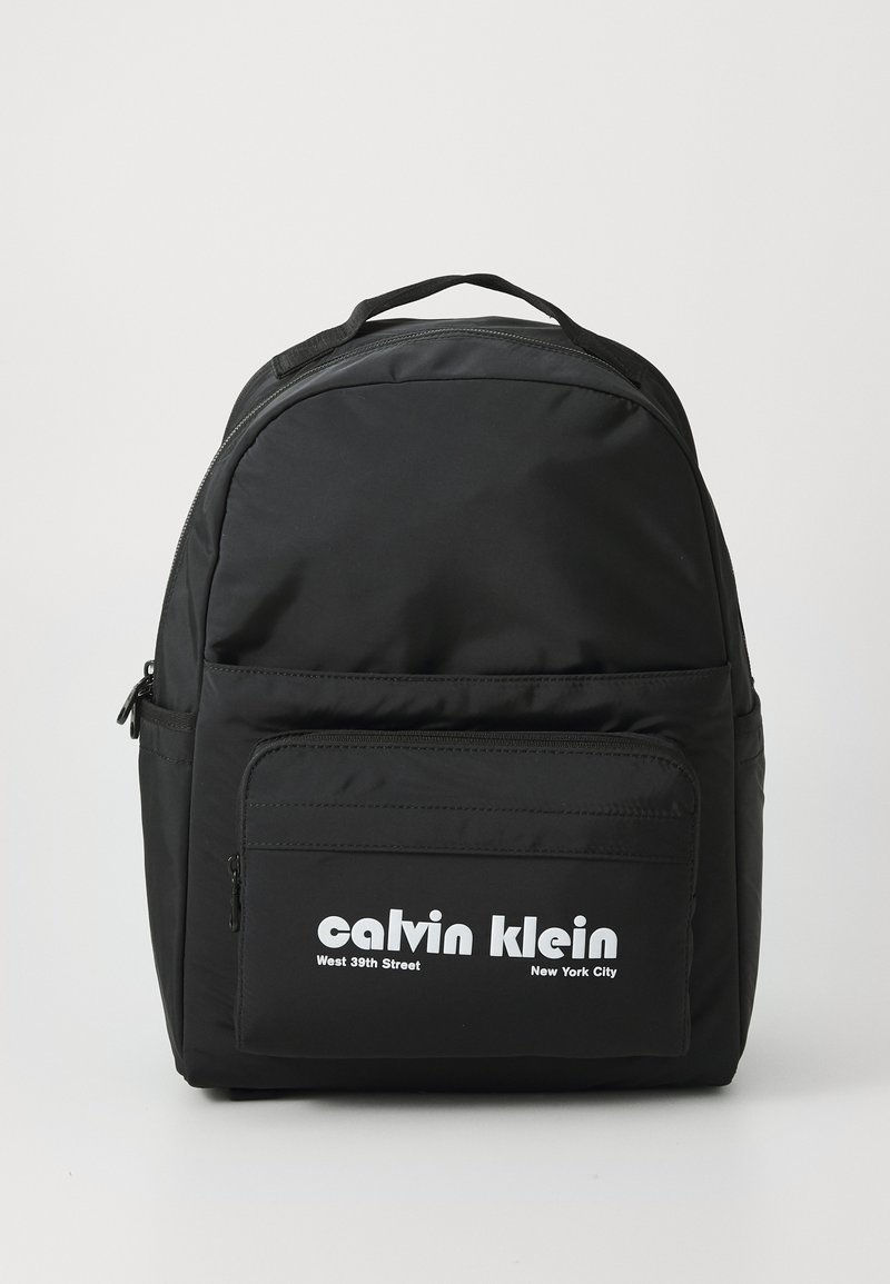 Sac à dos en nylon noir avec une forme arrondie, poche zippée à l'avant, marquage blanc proéminent et dos rembourré pour le confort.