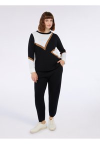 Maglione nero con design geometrici bianchi e marroni, abbinato a pantaloni neri e scarpe da ginnastica color crema. Tessuto morbido, vestibilità rilassata.