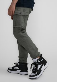 Groene cargobroek met voorzakken, gecombineerd met zwart-witte high-top sneakers met logo-accenten en textuurmateriaal.
