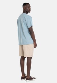 Camiseta de algodón azul claro de mangas cortas, combinada con shorts beige. El modelo lleva zapatillas deportivas marrón oscuro. Ajuste simple y relajado.