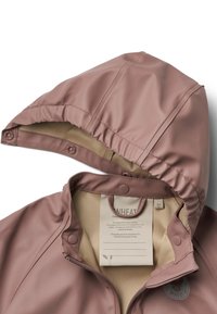 Vaaleanpunainen vedenpitävä takki hupulla, jossa on nepparit ja beige vuori. Sisäpuolella on hoito-ohjeet ja bränditiedot.