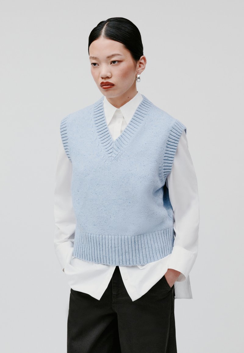 Lichtblauwe gebreide vest met een V-hals en ribbelrand, gedragen over een witte blouse met kraag. Soepele textuur, zonder mouwen.