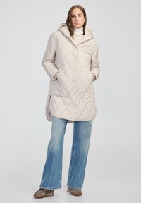 Béžový prošívaný dlouhý kabát s kapucí, s diagon�álním diamantovým vzorem a postranními kapsami, v kombinaci se světle modrými rozšířenými džíny a šedými botami.