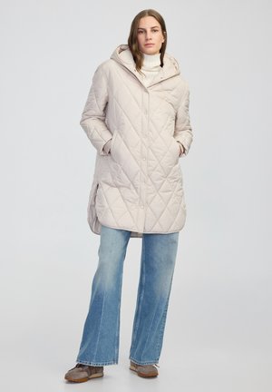 Beżowy pikowany długi płaszcz z kapturem, z ukośnym wzorem diamentów i bocznymi kieszeniami, w połączeniu z jasnoniebieskimi dzwonami i szarymi butami.