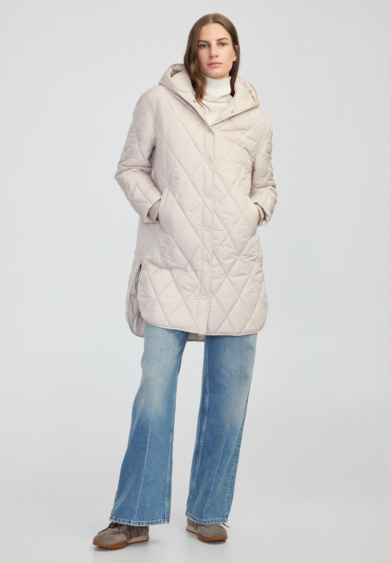 Béžový prošívaný dlouhý kabát s kapucí, s diagonálním diamantovým vzorem a postranními kapsami, v kombinaci se světle modrými rozšířenými džíny a šedými botami.