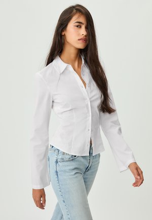 Weißes, tailliertes Hemd mit langen Ärmeln, spitzem Kragen und Knopfverschluss. Kombiniert mit hellblauen, hoch geschnittenen Jeans. Glatte Textur.