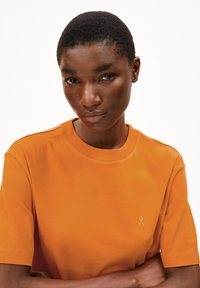 T-shirt en coton orange avec des manches courtes, un col rond et un logo brodé discret sur la poitrine. Texture lisse, coupe décontractée.