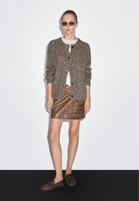 Frau steht und trägt eine braune Strickjacke, ein weißes Oberteil, einen braunen Leder-Minirock, dunkle Loafers und eine Sonnenbrille vor einem schlichten Hintergrund.