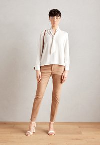 Weiße Bluse mit langen Ärmeln und Bindeband, beige taillierte Hose und weiße Sandalen mit Absatz. Das Outfit zeichnet sich durch ein minimalistisches Design und einen lässigen Look aus.