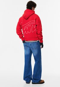 Punainen huppari, jossa valkoinen graafinen kuvio takana, yhdistettynä väljiin sinisiin farkkuihin ja ruskeisiin liukusomisteisiin kenkiin. Kankaan tekstuuri on näkyvissä.